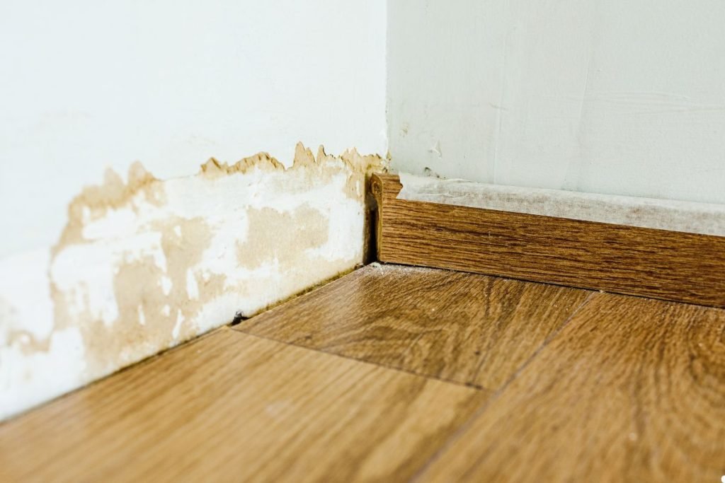 Water-damaged wall corner with peeling paint and stained drywall, alongside a wooden floor and baseboard.