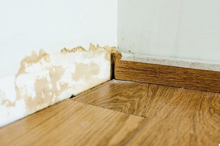 Water-damaged wall corner with peeling paint and stained drywall, alongside a wooden floor and baseboard.